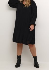 Robe noire oversize en tissu texturé, à manches longues et avec un petit décolleté en V. Portée avec des bottes noires hauteur cheville.