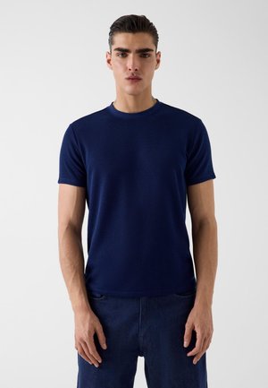 Homme debout de face portant une chemise ajustée bleu marine à manches courtes et un pantalon bleu foncé sur un fond clair uni.