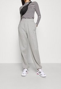 Persona vistiendo una camiseta de manga larga a rayas en blanco y negro, pantalones de chándal grises, zapatillas blancas y un bolso cruzado negro, contra un fondo liso.