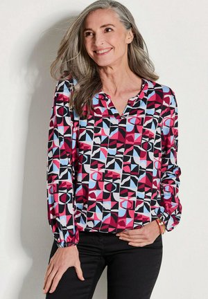 Langärmlige Bluse mit einem geometrischen Muster in Rot, Pink, Blau und Schwarz, V-Ausschnitt und gerafften Bündchen. Schwarze Hosen werden getragen.