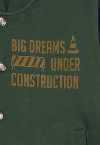 Grüne Stoffbluse mit silbernem Knopf, versehen mit gelbem Text "GROßE TRÄUME IM AUFBAU" und einer Grafik eines Baustellenkegels.