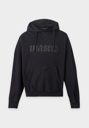 Мъж, гледащ напред, облечен в черен суичър с качулка с текст "wasted" и широки светлосини износени дънки с графичен принт отстрани.