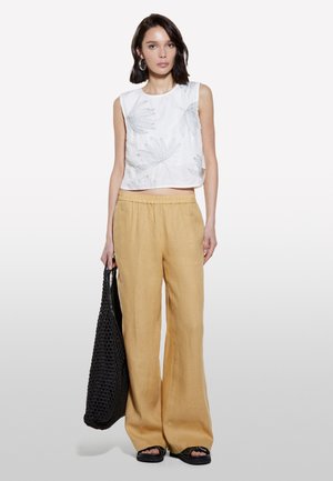 Haut court sans manches blanc avec impression de feuilles, associé à un pantalon large jaune moutarde. Le modèle tient un sac fourre-tout noir en tressé et porte des sandales noires.