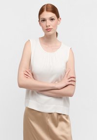 Mouwloze crèmekleurige blouse met een gathered halslijn en een zachte textuur; gecombineerd met een glanzende beige rok. Eenvoudige, casual fit.