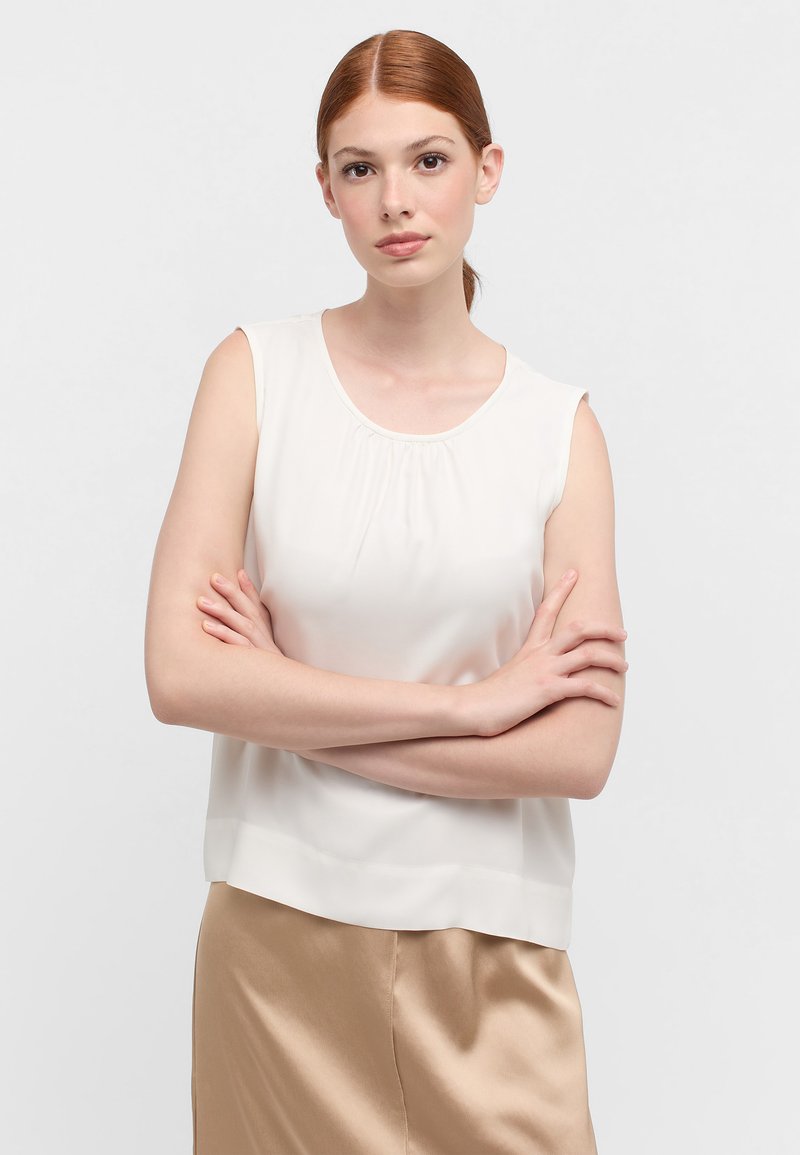 Mouwloze crèmekleurige blouse met een gathered halslijn en een zachte textuur; gecombineerd met een glanzende beige rok. Eenvoudige, casual fit.