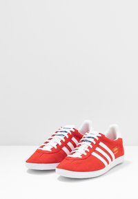 Röda mockaskor med vita gummisulate, med tre vita ränder, blå snören och en gul logga på sidan.