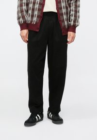 Cardigan à carreaux bordeaux et gris avec fermeture à boutons, associé à un pantalon noir ample et à des baskets noires avec des bandes blanches.