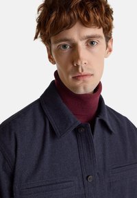 Camisa chaqueta de mezcla de lana azul marino con cierre de botones, dos bolsillos en el pecho y cuello con solapa, llevada sobre un jersey de cuello alto burdeos.
