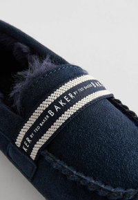 Mocassino in suede blu navy con fascia a righe bianche e blu navy. Presenta una fodera morbida e cuciture decorative lungo il bordo.