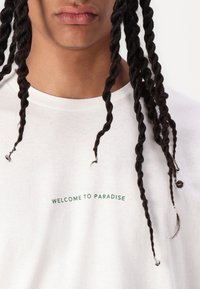 Hvid bomulds t-shirt med grøn tekst, der står "VELKOMMEN TIL PARADIS." Vist med fletning af mørkt hår i forgrunden.