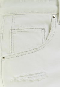 Tejido de mezclilla blanca con detalles desgastados, un pequeño bolsillo trasero, costuras en contraste y un remache de metal plateado.
