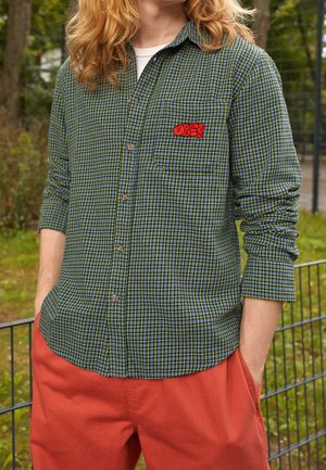 Personne portant une chemise à manches longues à carreaux verts et bleus avec un patch rouge "OBEY" sur la poche et un pantalon rouge ample, debout à l'extérieur près d'une clôture métallique.