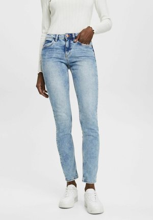 Frau trägt hellblaue Skinny Jeans, weißes geripptes Langarmshirt und weiße Sneakers, steht mit einer Hand in der Tasche.