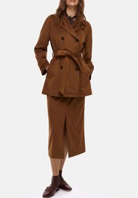 Trench coat doppio petto marrone con cintura, abbinato a una gonna coordinata al ginocchio con uno spacco frontale. Gli accessori includono scarpe marroni.