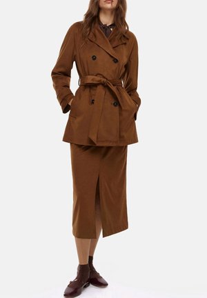 Trench coat doppio petto marrone con cintura, abbinato a una gonna coordinata al ginocchio con uno spacco frontale. Gli accessori includono scarpe marroni.