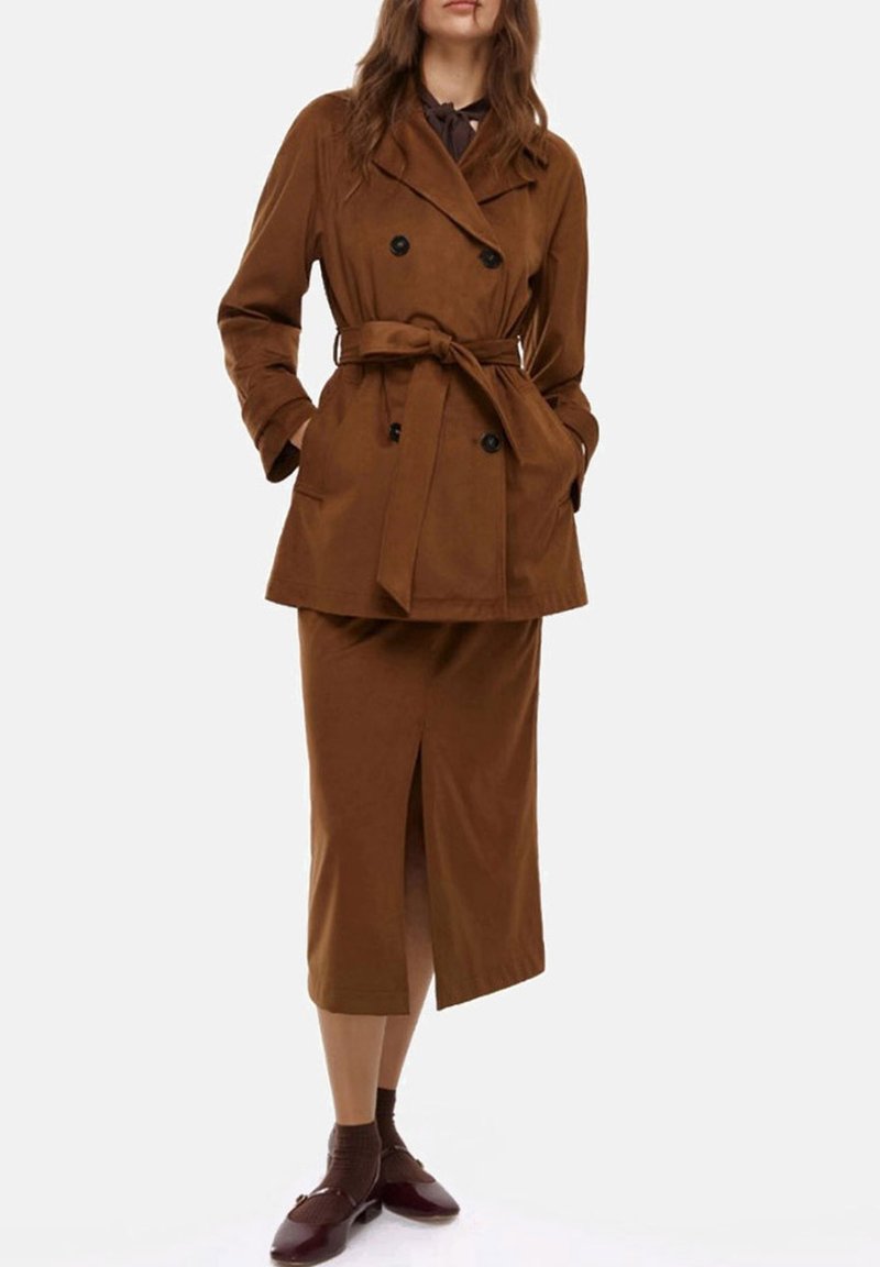 Trench coat doppio petto marrone con cintura, abbinato a una gonna coordinata al ginocchio con uno spacco frontale. Gli accessori includono scarpe marroni.