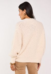 Maglione lavorato a maglia color crema con una vestibilità ampia, texture a coste e maniche larghe. Indossato sopra pantaloni in velluto a coste marrone chiaro, con dettagli in oro sui bracciali.