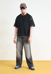 Chemise noire à manches courtes avec des accents gris, associée à un jean gris et des chaussures à accents bleus. Le modèle porte une casquette marron et des lunettes de soleil noires.
