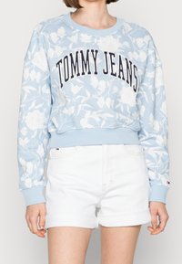 Světle modrá květinová mikina s bílými květinovými vzory, žebrované manžety a výstřih. Na hrudi se nachází nápis "TOMMY JEANS" v tučném námořnickém písmu.