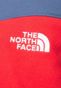 Il tessuto in pile rosso e blu presenta un logo ricamato in bianco de "THE NORTH FACE" con un design a linee curve. La texture è morbida e calda.