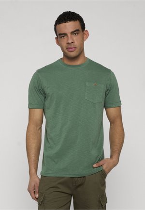 Homme aux cheveux bouclés courts portant un T-shirt vert à manches courtes avec poche poitrine et un pantalon cargo olive, debout devant un fond gris.