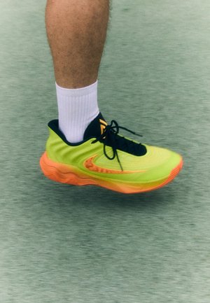 Jambe portant une chaussure de course vert fluo et orange avec des lacets noirs et une chaussette blanche sur une surface texturée grise.