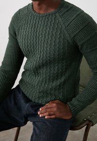 Groen gebreide trui met een gestructureerd kabelpatroon, ronde halslijn en aangesloten mouwen, gecombineerd met donkere spijkerbroek.