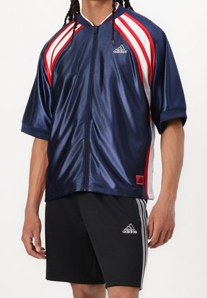Personne portant un haut de sport zippé Adidas bleu marine avec des rayures rouges et blanches et un short Adidas noir avec des rayures blanches.