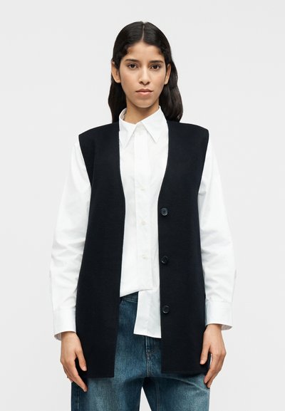Gilet blu navy lavorato a maglia con vestibilità rilassata, senza maniche, scollo rotondo e tre bottoni, indossato sopra una camicia bianca.