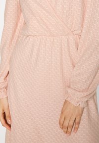 Vestido rosa claro com material texturizado, mangas longas e punhos elásticos. Apresenta um design envolvente e uma cintura franzida para um visual ajustado.