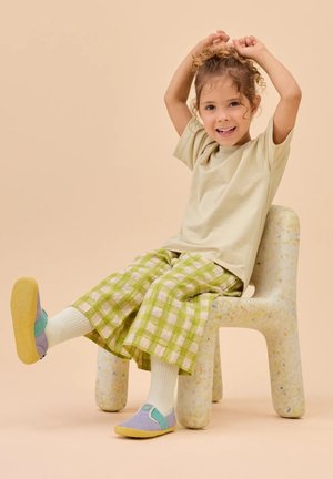 Hellbeiges T-Shirt, grüne, karierte, weit geschnittene Hose, lila-türkisfarbene Schuhe, weiße Sportsocken, sitzend auf einem gesprenkelten, abgerundeten Stuhl.