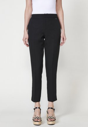 Pantalons noirs ajustés avec une coupe droite, présentant une finition en tissu lisse et des détails discrets sur les coutures latérales, associés à des sandales à plateformes noires et blanches imposantes.