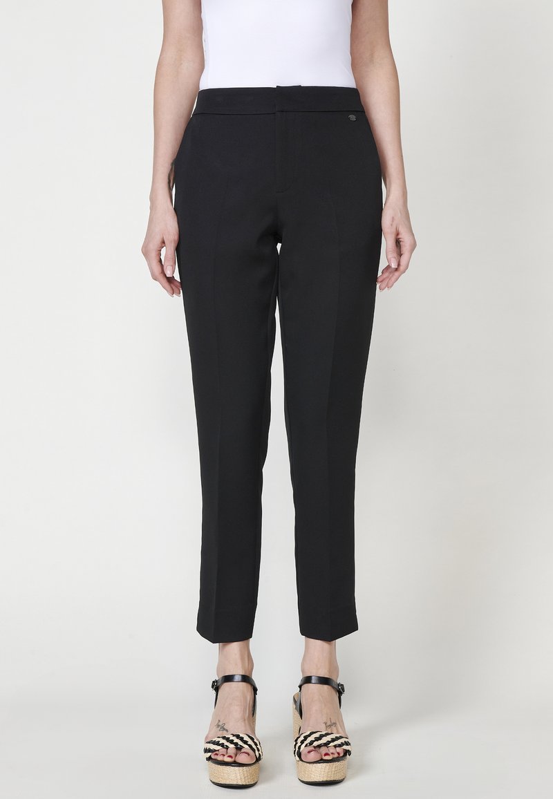 Pantalons noirs ajustés avec une coupe droite, présentant une finition en tissu lisse et des détails discrets sur les coutures latérales, associés à des sandales à plateformes noires et blanches imposantes.
