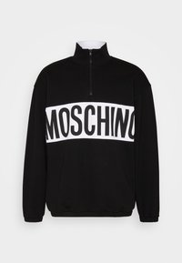 Černý svetr s vysokým límcem a zipem, zdobený bílým pruhem přes hrudník s nápisem "MOSCHINO" tučným černým písmem. Měkká textilie.
