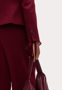 Blazer ajustado burdeos con manga y detalle de botón negro, combinado con pantalones a juego; mano sosteniendo un bolso de cuero burdeos con cremallera.
