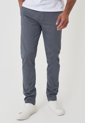 Mann trägt gerade geschnittene graue Hose und weiße Turnschuhe und steht mit einer Hand entspannt an seiner Seite vor einem einfarbigen Hintergrund.