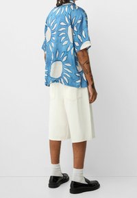 Niebieska koszula z krótkim rękawem w wzory słoneczne, zestawiona z białymi szortami dżinsowymi. Czarne buty, widoczne białe skarpetki. Szczegółowy tatuaż na lewej ręce.