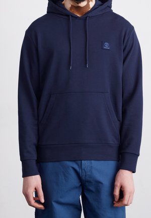 Mies pukeutunut tummansiniseen Timberland-huppariin, jossa etutasku ja kiristysnauhat, yhdistetty sinisiin housuihin, kuvattuna kaulasta lantioon.