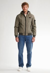 Giacca bomber verde oliva con cappuccio, tasche con zip e polsini a costine, abbinata a jeans blu chiaro e sneakers bianche.