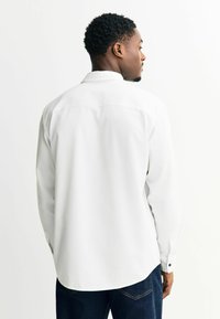 Hvid langærmet skjorte med en struktureret overflade, spids krave og knapmanchetter, set bagfra, parret med mørkeblå jeans.