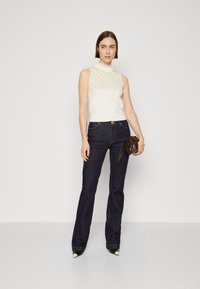 Mouwloze crèmekleurige top met een gestippeld kantpaneel, gecombineerd met donkerblauwe flared jeans met gouden knoopaccenten en een bruine handtas.