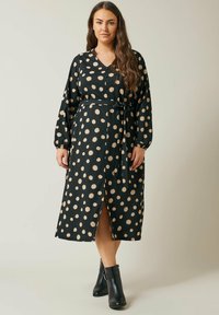 Lang schwarzes Kleid mit cremefarbenen Punkten, V-Ausschnitt, langen Ärmeln, einem Taillenband und einem Schlitz vorne. Aus strukturiertem Stoff gefertigt. Schwarze Stiefeletten.