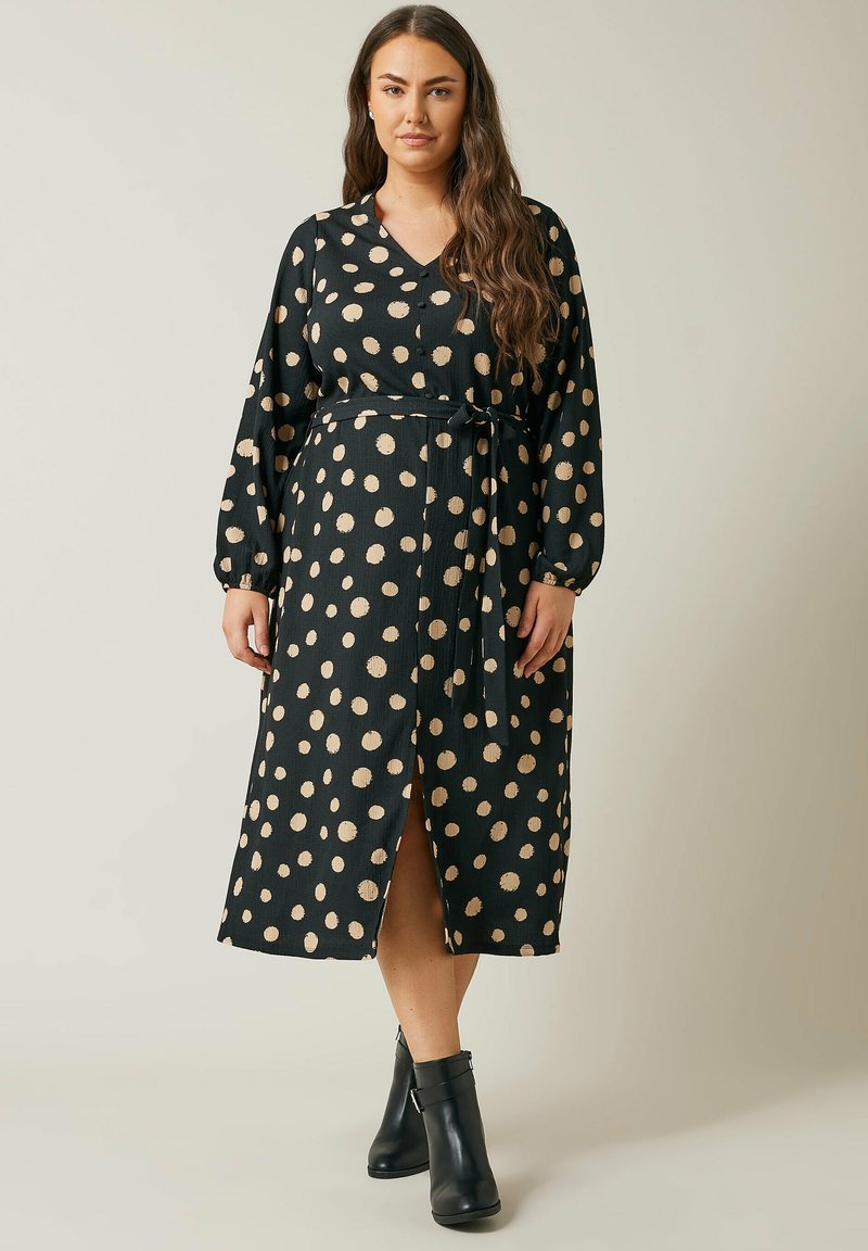Lang schwarzes Kleid mit cremefarbenen Punkten, V-Ausschnitt, langen Ärmeln, einem Taillenband und einem Schlitz vorne. Aus strukturiertem Stoff gefertigt. Schwarze Stiefeletten.