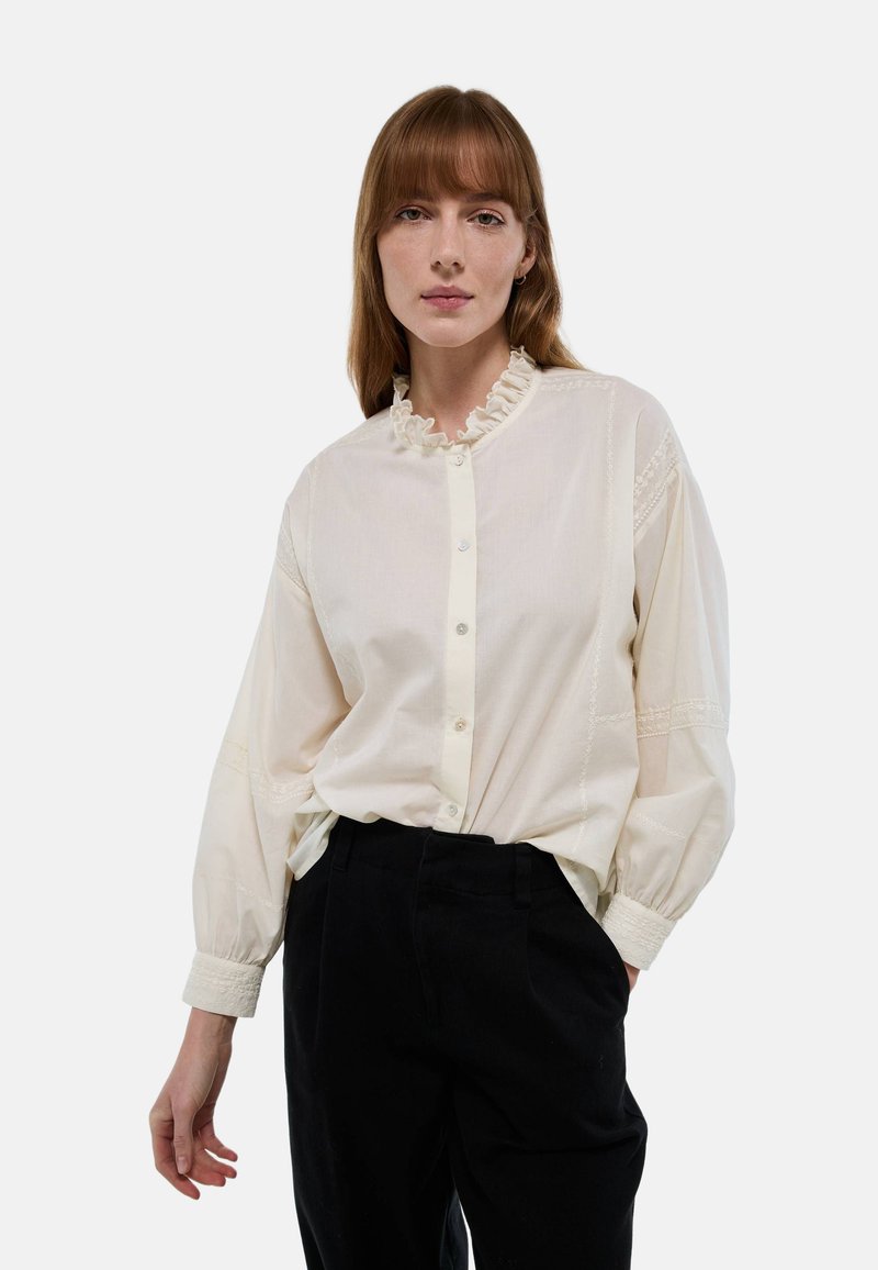 Blouse blanche avec col volants et fermeture à boutons, présentant des coutures décoratives et des manches longues. Portée avec un pantalon noir taille haute.