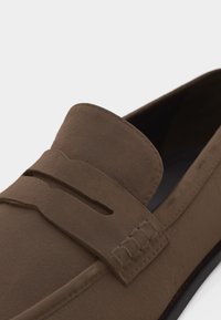 Mocassin en daim marron avec un bout mocassin et une bride décorative sur le dessus.