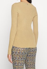 Goldfarbener, figurbetonter Pullover mit langen Ärmeln und strukturierter Oberfläche sowie rundem Halsausschnitt. Mit einem kleinen Detail am Rücken am Halsausschnitt. Kombiniert mit gemusterter Hose.