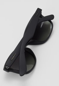 Lunettes de soleil en plastique noir avec des verres teintés, présentant une finition mate lisse et un logo sur la branche. Forme unique avec des branches courbées.
