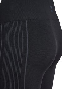 Schwarze Leggings mit einer strukturierten, gerippten Oberfläche auf einer Seite, gefertigt aus elastischem Material mit hohem Bund. Dezente Markenlogos sind sichtbar.