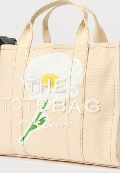 Béžová plátenná taška s veľkým bielym dizajnom sedmokrásky, vyšitým textom "THE TOTE BAG" a mäkkými rúčkami na nosenie.