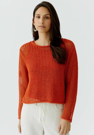 Vrouw met lang donker haar die een feloranje opengewerkte trui draagt over een rood shirt en witte broek, frontaal kijkend tegen een effen achtergrond.